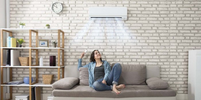 Eine Junge Frau sitzt in einem Raum auf der Couch mit der Fernbedienung für die Klimaanlage die an der Wand über Ihr ist. Aus der Klimaanlage kommt kühle Luft, und die Frau atmet Tief und lächelt.