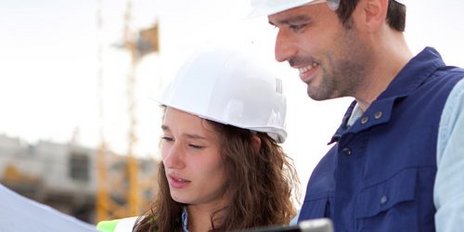 Eine Frau mit Bauhelm hält eine Zeichnung neben ihr steht ein Mann mit einem Tablet auf einer Baustelle